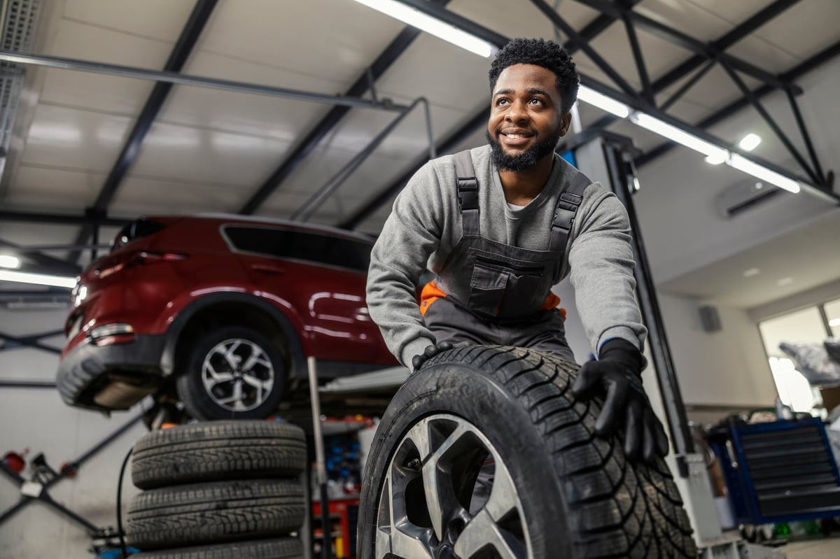 Hardworking smiling car technician rolling a car tyre towards the camera at an automotive service center
